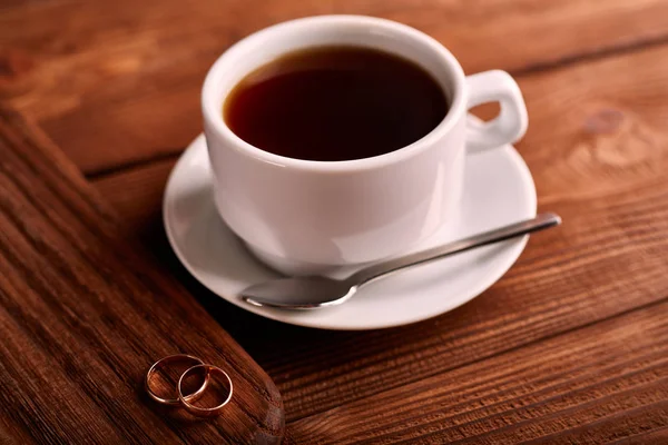Preto, sabor, café orgânico em copo clássico branco em uma mesa de madeira com um par de alianças de casamento . — Fotografia de Stock