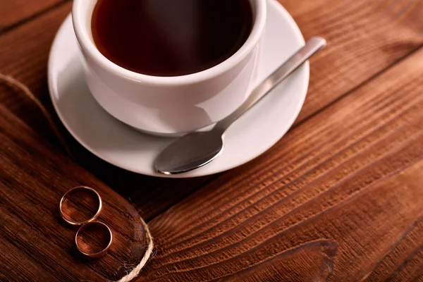 Preto, sabor, café orgânico em copo clássico branco em uma mesa de madeira com um par de alianças de casamento . — Fotografia de Stock