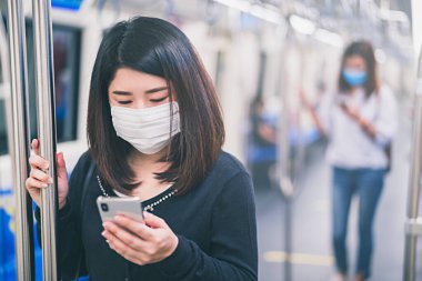 Korumalı yüz maskesi takan Asyalı genç bir kadın, kirli hava veya 22.5 ve Coronavirüs veya COVID-19 salgını nedeniyle yeraltı trenlerinde akıllı telefon kullanıyor..