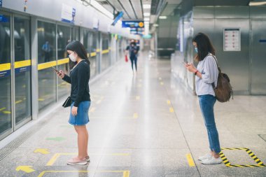 Korumalı yüz maskesi takan iki genç Asyalı kadın, metroda Coronavirus veya COVID-19 salgını nedeniyle dünyanın tüm karasal kesimlerinde metroda bekleme sırasında sosyal mesafeye göre sırada bekliyor.