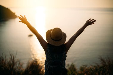 Sırt çantalı ve şapkalı Asyalı genç bir gezgin Promthep Cape Phuket Tayland 'da gün batımını izlerken iki elini mutlulukla kaldırıyor. Özgürlük mutluluk komik bir kavram