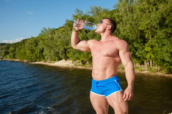 Schöner, muskulöser Mann trinkt Wasser am Strand — Stockfoto