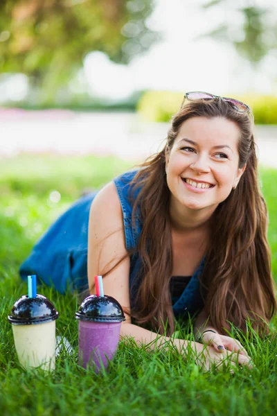 Молодая женщина в солнечных очках пьет сочные вкусные молочные коктейли на зеленой газонной траве — стоковое фото