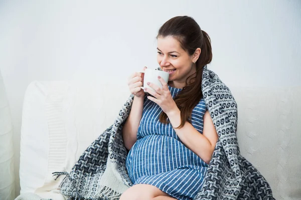 Беременная женщина пьет чай и под теплым уютным пледом дома — стоковое фото