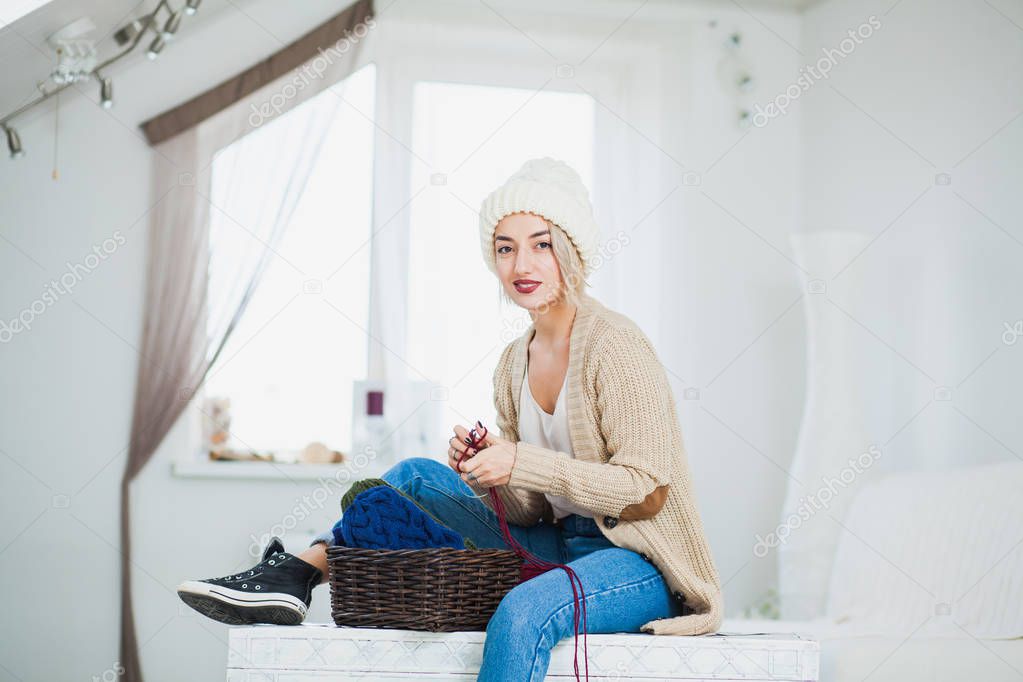 Beautiful woman in big white knitted hat indoors sit on chest with basket yarn. Knitting at home, hobby, cozy flat, denim casual clothing.