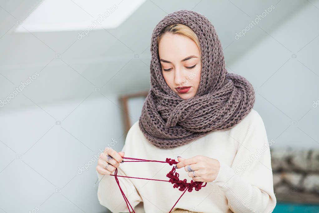 Young woman in warm hand knitted snood scarf at home. Beautiful girl knitting in cozy flat wearing casual clothes, big brown scarf.