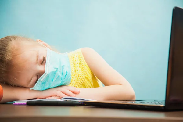 Antiviral maskeli küçük bir ilkokul kızı laptopun yanında uyuyor..