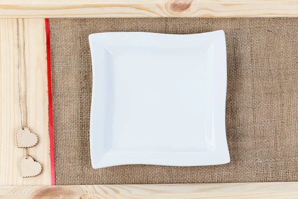 Heart on a background of wood covered with burlap, a white square plate a card for Valentine's Day. — Stock Photo, Image