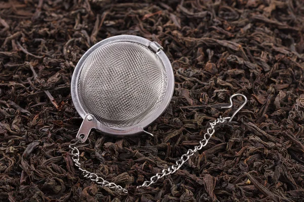 A tea filter on a chain with black tea on a pile of dry tea leaves. A tea filter is a close-up of a kitchen accessory.