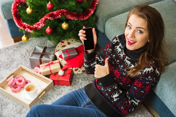 Joven mujer sonriente sosteniendo el teléfono y mostrando el pulgar hacia arriba en Navidad — Foto de Stock