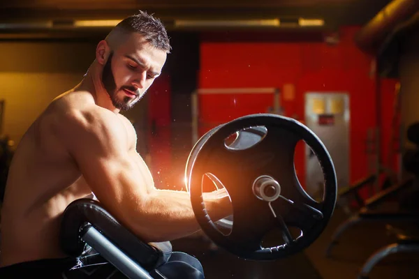 Jovem musculoso homem formação com sinos e olhando para bíceps — Fotografia de Stock