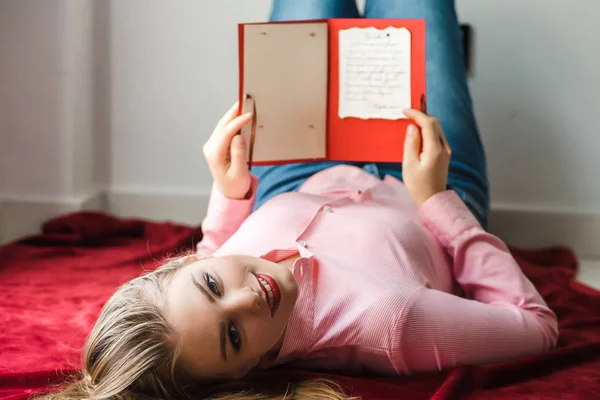 Joven chica rubia sonriente con el pelo largo acostado en la cama y sosteniendo la tarjeta hecha a mano en sus manos —  Fotos de Stock