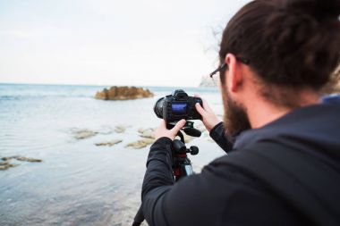 Genç yaratıcı fotoğrafçı bir tripod üzerinde kamera ile deniz ve kayalar fotoğraflarını yapma gözlük.