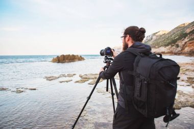 Sakal ve bir tripod üzerinde kamera ile deniz ve kayalar fotoğraflarını yapım sırt çantası ile genç şık fotoğrafçı. Akdeniz