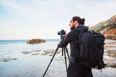 Sakal ve bir tripod üzerinde kamera ile deniz ve kayalar fotoğraflarını yapım sırt çantası ile genç şık fotoğrafçı. Akdeniz