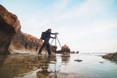 Sakal ve bir tripod üzerinde kamera ile deniz ve kayalar fotoğraflarını yapım sırt çantası ile genç şık fotoğrafçı. Akdeniz