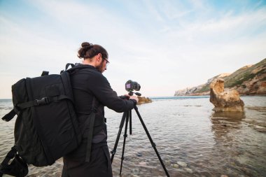 Sakal ve bir tripod üzerinde kamera ile deniz ve kayalar fotoğraflarını yapım sırt çantası ile genç şık fotoğrafçı. Eğer görünüm şaşırtıcı Akdeniz deniz ve gökyüzü