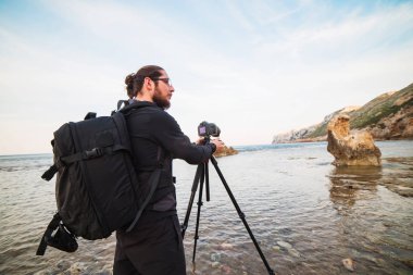 Sakal ve bir tripod üzerinde kamera ile deniz ve kayalar fotoğraflarını yapım sırt çantası ile genç şık fotoğrafçı. Akdeniz