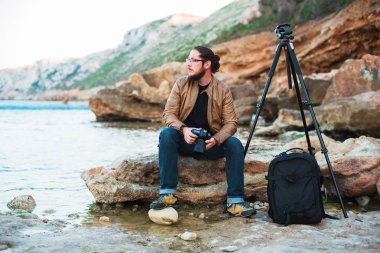 Genç şık fotoğraf sakallı ve kayanın üzerinde oturan ve denizde seyir elinde fotoğraf makinesi ile gözlük. Tripod ve onun yanında sırt çantası.