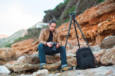 Gözlük kayanın üzerinde oturan ve onun kamera üzerinde yaptığı fotoğraf izlerken sakallı genç şık fotoğrafçıymış. Tripod ve onun yanında sırt çantası.