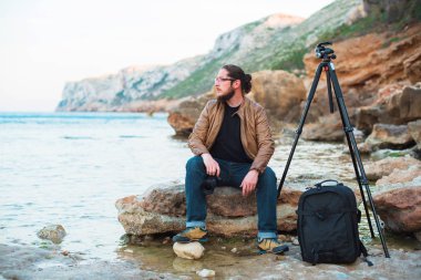 Genç şık fotoğraf sakallı ve kayanın üzerinde oturan ve denizde seyir elinde fotoğraf makinesi ile gözlük. Tripod ve onun yanında sırt çantası.