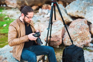 Gözlük kayanın üzerinde oturan ve onun kamera üzerinde yaptığı fotoğraf izlerken sakallı genç şık fotoğrafçıymış. Tripod ve onun yanında sırt çantası.