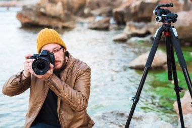 Renksizdir sarı kap ve kayanın üzerinde oturan ve fotoğrafın yapma gözlük sakallı genç şık fotoğrafçı. Tripod ve sırt çantası onun yanında. Sıcak deniz