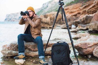 Renksizdir sarı kap ve kayanın üzerinde oturan ve deniz fotoğraf yapma gözlük sakallı genç şık fotoğrafçı. Tripod ve onun yanında sırt çantası.