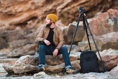 Sarı kap ve onun elinde profesyonel kamera ile kayaya oturan ve denize bakarak gözlük renksizdir sakallı genç şık fotoğrafçı. Tripod ve onun yanında sırt çantası.