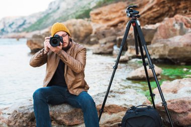 Renksizdir sarı kap ve kayanın üzerinde oturan ve fotoğrafın yapma gözlük sakallı genç şık fotoğrafçı. Tripod ve sırt çantası onun yanında. Sıcak deniz