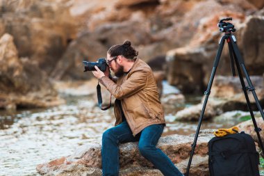 Kayanın üzerinde oturan ve deniz fotoğraf yapma uzun saç ve sakal ile genç şık fotoğrafçı. Tripod ve onun yanında sırt çantası.