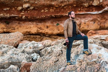 Sakallı, uzun saç ve onun elinde fotoğraf makinesi ile bir rock kumsalda ayakta ve mesafe içine bakarak rahat kıyafetler giyen gözlük ile genç erkek fotoğraf.