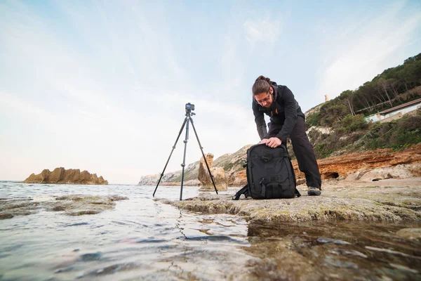 年轻的时尚摄影师与胡子寻找东西在他的专业背包 相机在三脚架上 地中海 — 图库照片