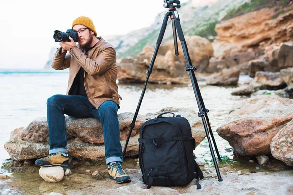 Renksizdir sarı kap ve kayanın üzerinde oturan ve deniz fotoğraf yapma gözlük sakallı genç şık fotoğrafçı. Tripod ve onun yanında sırt çantası.