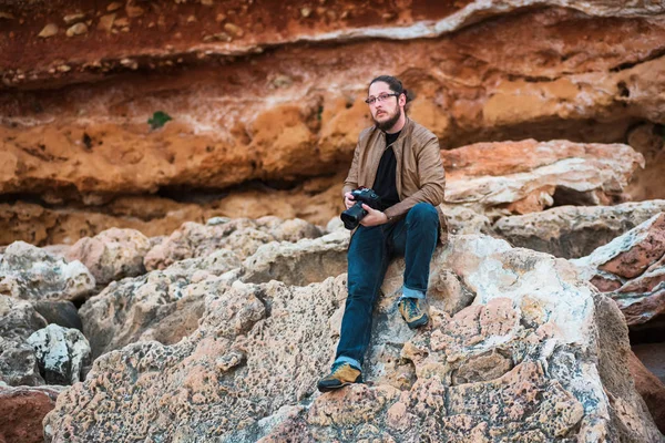 Young male photographer with beard, long hair and glasses wearing casual clothes sitting on a rock beach with camera in his hands and looking into the distance.