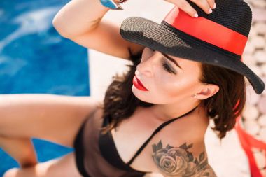 Genç seksi moda kadın siyah şapka ve mayo kırmızı dudaklı yüzme havuzunda poz. Takı, dövme, parlak makyaj. Moda stil. Yaz ve tatil kavramı.