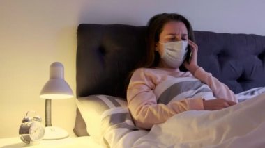 Yüzünde tıbbi maske olan hasta bir kadın evde yatakta yatıyor, öksürüyor ve doktor çağırıyor. Koronavirüs belirtileri gösteren bir kadın. Covid-19 enfeksiyonu.