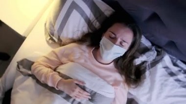 Sağlık maskeli genç bir kadının evde yatarken korkunç bir baş ağrısı çekmesi. Coronavirus belirtileri. Vertigo yakınlaştırması.