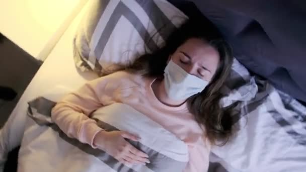 自宅で寝そべっている間にひどい頭痛に苦しむ医療用マスクの若い女性のトップビュー コロナウイルスの症状 めまいズーム — ストック動画