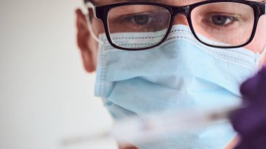 Gözlüklü, tıbbi maskeli ve tek kullanımlık eldivenli genç adam beyaz arka planda izole edilmiş termometreye bakıyor. Coronavirus, Covid-19, 2019-NCoV salgını. Karantina, evde kal. Boşluğu kopyala.