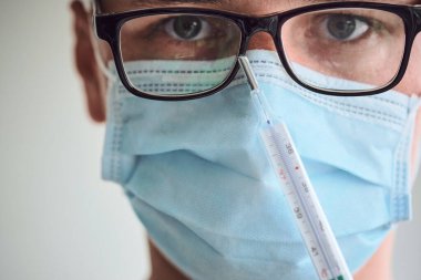 Gözlüklü, tıbbi maskeli ve termometreyi elinde tutan tek kullanımlık eldivenli genç adamın yakın plan portresi. Coronavirus, Covid-19, 2019-NCoV salgını. Boşluğu kopyala.