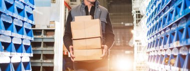 Delivery man in gray uniform holds boxes in his hands at the warehouse. Gold backlight, widescreen image. clipart