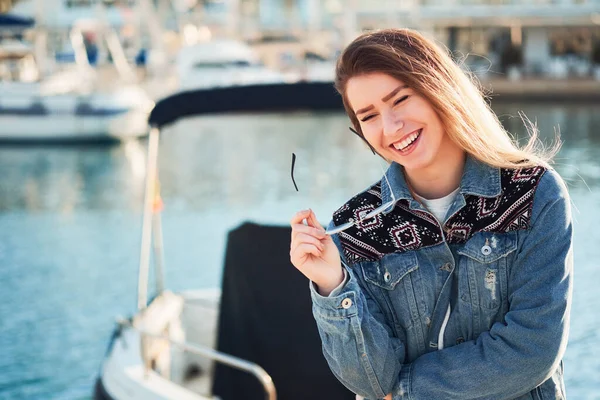 Beautiful girl naturally laughs and holds round glasses in her hand with blond hair on a summer day outdoors in the port