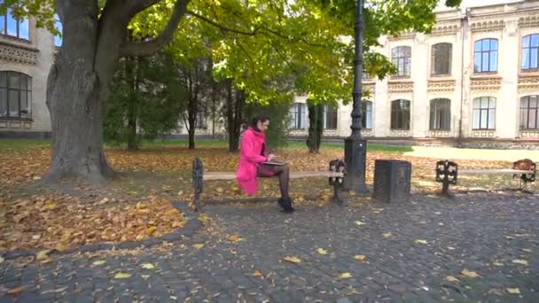 Giovane ragazza in cappotto rosa con computer portatile su panchina nel parco universitario — Video Stock