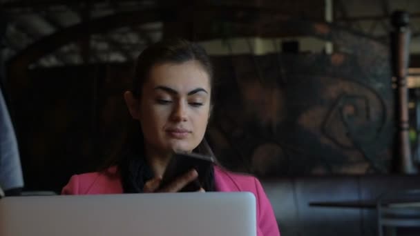 Молодая девушка с ноутбуком за столом в кафе разговаривает по телефону — стоковое видео