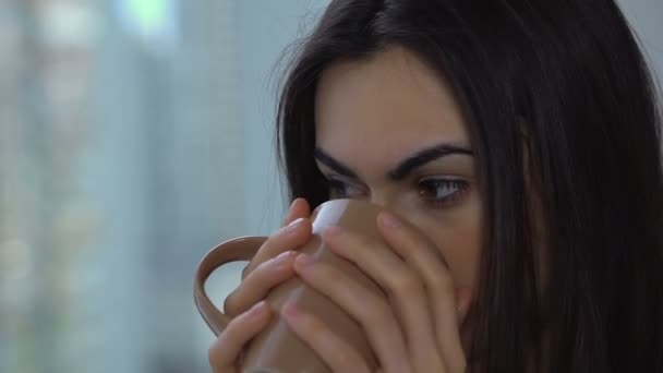 Красива брюнетка п'є каву і посміхається дивитися у вікно крупним планом — стокове відео