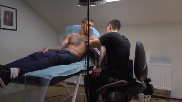 Tattoo workshop client met grimas op zijn gezicht iets last heeft van pijn tijdens het maken van een tatoeage — Stockvideo