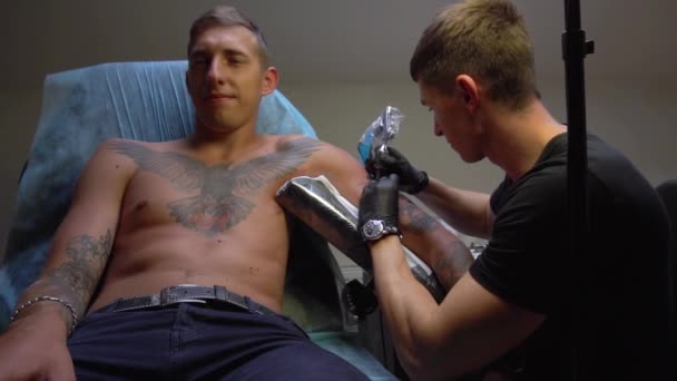 Uśmiecha się facet siedzi w fotelu, podczas gdy kapitan sprawia mu tatuaż na ramieniu — Wideo stockowe