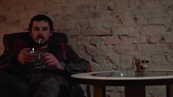 L'uomo accende la sigaretta e mette l'accendino sul tavolo accanto al vetro e al cranio — Video Stock
