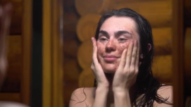 Menina morena após sauna feliz e sorrindo aplica esfrega de café em seu rosto olhando no espelho — Vídeo de Stock